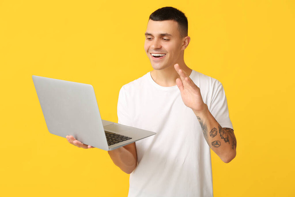 Beau jeune homme avec ordinateur portable agitant la main sur fond jaune - Photo, image