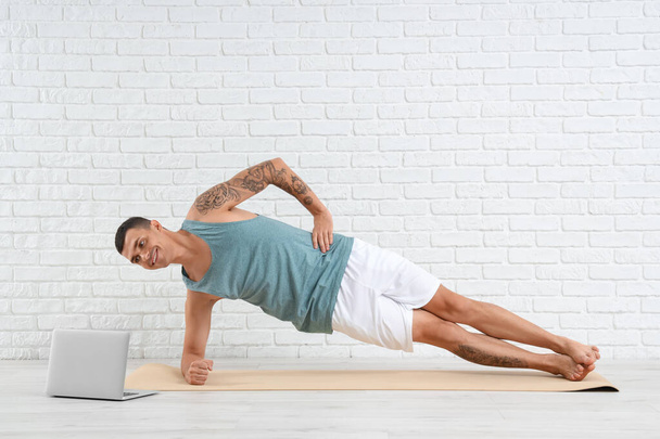 Молодой человек с ноутбуком делает йогу на коврике возле стены из белого кирпича - Фото, изображение