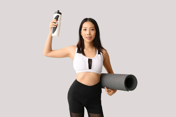 Sporty young Asian woman with water bottle and fitness mat on light background - Photo, Image