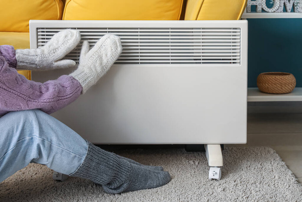 Mujer joven en cálidos calcetines de invierno y guantes cerca de calentador eléctrico. Concepto de temporada de calefacción - Foto, Imagen