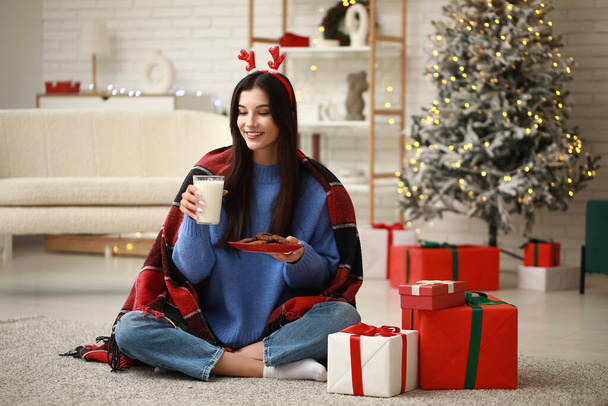 Bella giovane donna in corna di renna con bicchiere di latte e biscotti a casa la vigilia di Natale - Foto, immagini