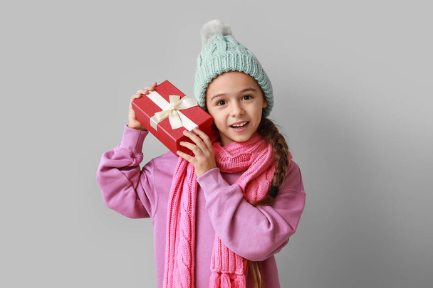 Χαριτωμένο κοριτσάκι σε ζεστό καπέλο και μαντήλι με χριστουγεννιάτικο δώρο σε γκρι φόντο - Φωτογραφία, εικόνα