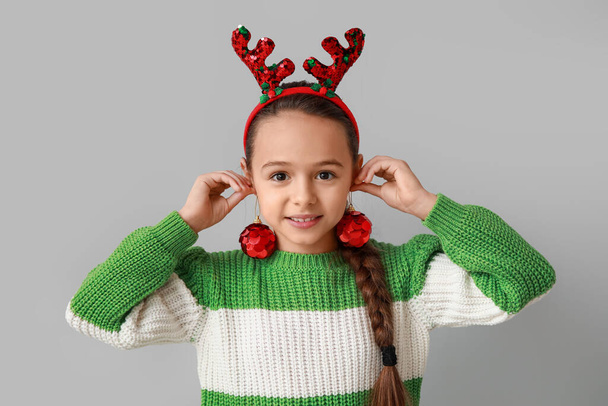 Nettes kleines Mädchen in Rentierhörnern mit Weihnachtskugeln auf grauem Hintergrund - Foto, Bild