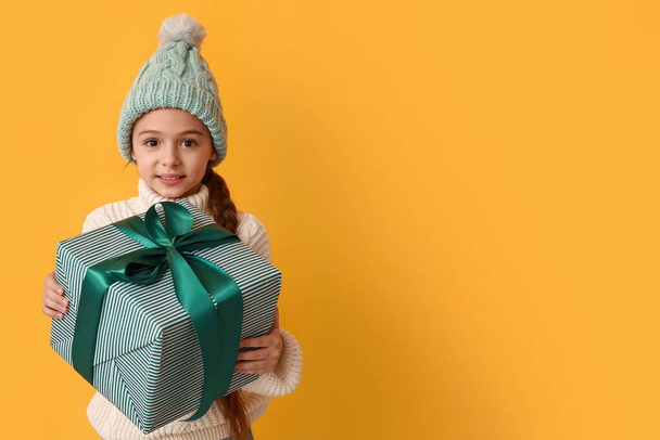 Schattig klein meisje met warme hoed met kerstcadeau op gele achtergrond - Foto, afbeelding