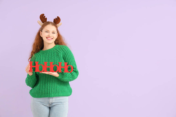 Young woman in reindeer horns with Christmas greeting on lilac background - Photo, Image