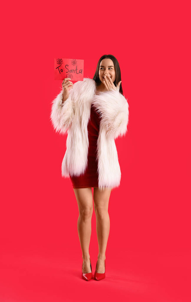 Surprised young woman with letter to Santa on red background - Photo, Image