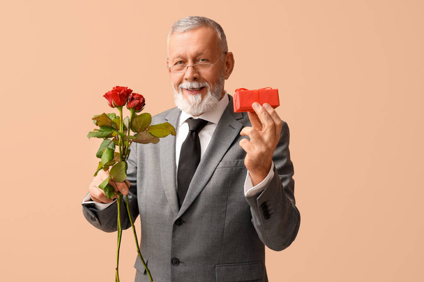 Dojrzały mężczyzna z różanymi kwiatami i prezentem na Walentynki na beżowym tle - Zdjęcie, obraz