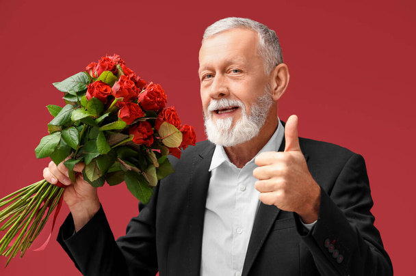 Älterer Mann mit einem Rosenstrauß, der den Daumen nach oben zeigt auf rotem Hintergrund. Valentinstag-Feier - Foto, Bild