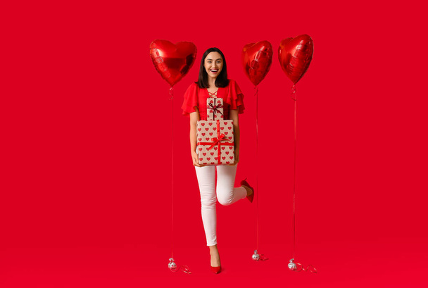 Kaunis nuori nainen, jolla on ilmapalloja sydämen ja lahjapakkausten muodossa punaisella taustalla. Ystävänpäivä juhla - Valokuva, kuva