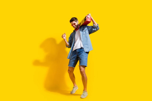 Foto corpo pieno di uomo eccentrico vestito pantaloncini camicia denim in occhiali danza con boombox sulla spalla isolato su sfondo di colore giallo. - Foto, immagini