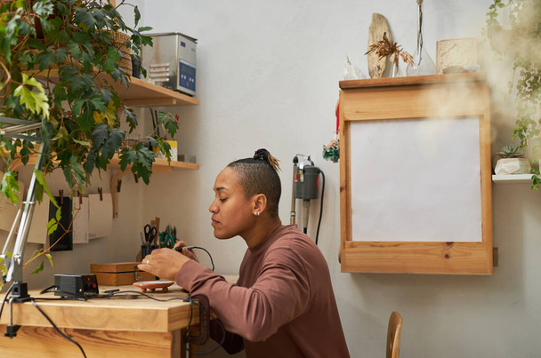 Retrato de vista lateral de artista femenina negra creando joyas hechas a mano en un acogedor taller, espacio para copiar - Foto, Imagen