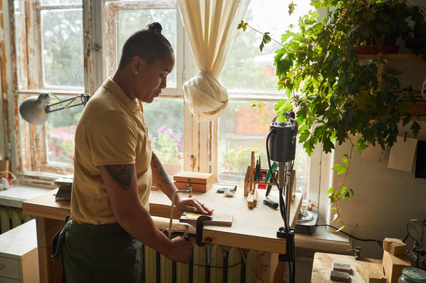 Porträt einer tätowierten Künstlerin, die Holz schneidet, während sie am Fenster in einer gemütlichen Werkstatt steht, Kopierraum - Foto, Bild