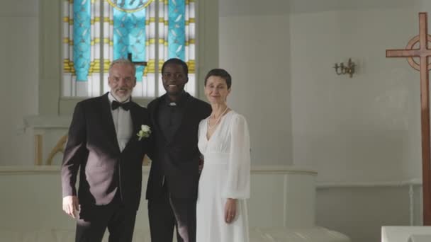 Retrato médio de casal recém-casado caucasiano maduro e seu oficiante masculino preto posando para câmera em pé na capela do casamento branco após a cerimônia - Filmagem, Vídeo