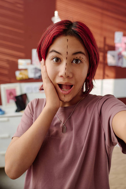 Ohromená teenager s výtahy na tváři dotýká její tvář při výrobě selfie nebo dělat video o plastické chirurgii pro předplatitele - Fotografie, Obrázek