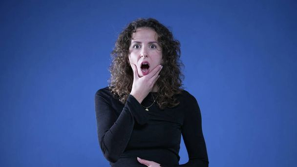 Überraschte Frau reagiert mit Ungläubigkeit, indem sie Mund mit der Hand bedeckt und in die Kamera auf blauem Hintergrund schaut - Foto, Bild