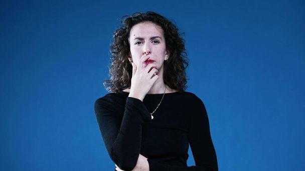 Kontemplative Entscheidungsfindung einer Frau beim Schielen und mit Hand in Kinn grübelnder Lösung, die auf blauem Hintergrund in die Ferne blickt - Foto, Bild