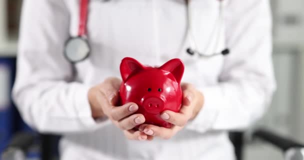 Woman doctor is holding piggy bank in hands. Treatment cost concept - Footage, Video