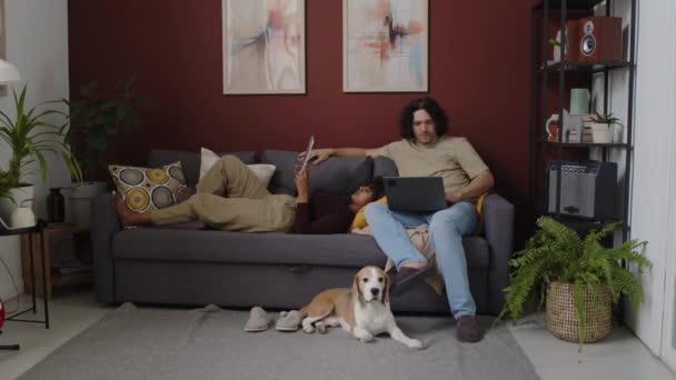 Lange Aufnahme eines jungen gemischtrassigen Paares mit kleinem Beagle-Hund, das es sich zu Hause auf dem Sofa gemütlich macht. Zweirassiges Mädchen liegt mit digitalem Tablet auf den Schenkeln ihrer kaukasischen Freunde und er surft am Laptop - Filmmaterial, Video