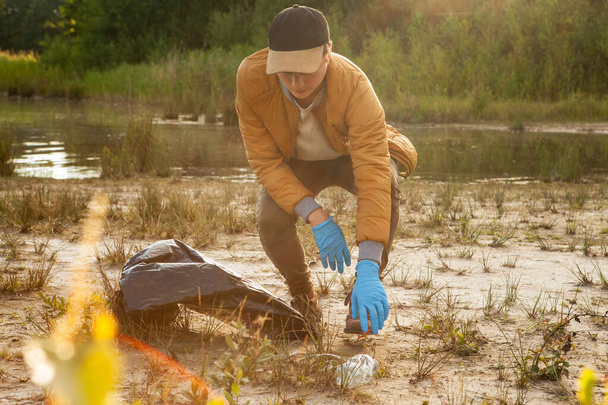 La foto captura a un solo voluntario, un joven caucásico, comprometido en un esfuerzo de conservación, limpiando una zona junto al lago. Se agachó para recoger la basura, usando guantes azules para proteger su - Foto, imagen