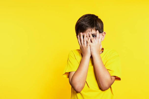 Kind in gelber Kleidung blickt durch Finger, die sein Gesicht bedecken, eine Geste der Neugier und Überraschung, vor einem monochromen gelben Hintergrund. - Foto, Bild