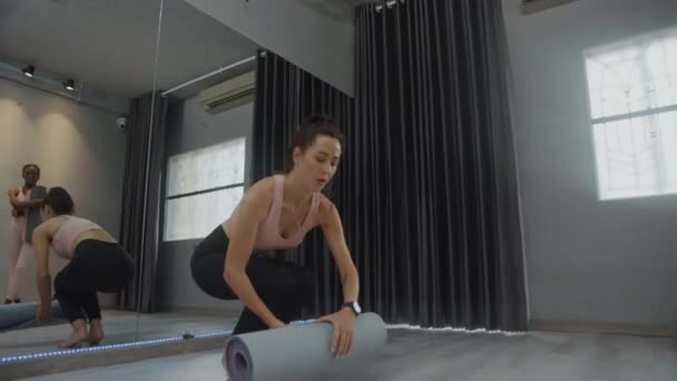 Stüdyoda diğer kadınlarla derse hazırlanırken genç yoga eğitmeninin minderi yuvarlarken çekilen görüntüsü. - Video, Çekim