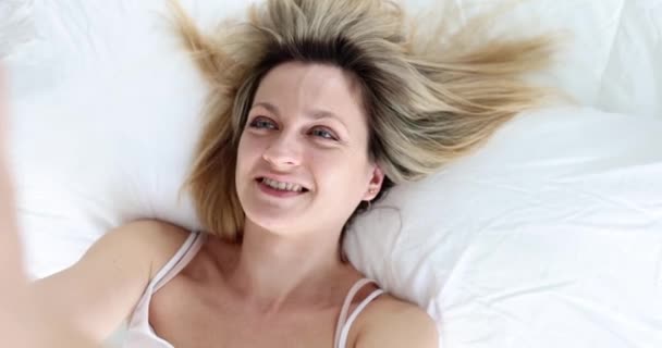 Mujer feliz se toma una selfie mientras está acostado en la cama, de cerca, tiro de mano. Personalidad de Internet, alegría del narcisismo - Metraje, vídeo
