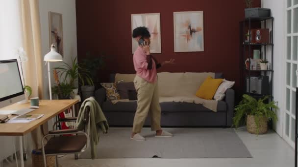 Volledig shot van de jonge Biracial vrouw lopen heen en weer in de stijlvolle woonkamer tijdens het gesprek op de smartphone - Video