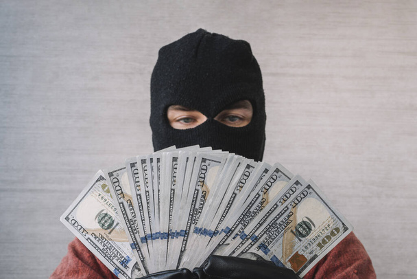 Homem ladrão com balaclava na cabeça segurando um punhado de notas de dólar na frente dos olhos. o conceito de crime e roubo de dinheiro - Foto, Imagem
