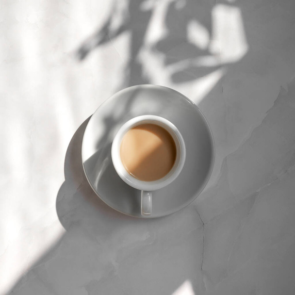 Минималистская эстетическая концепция доброго утра, белая керамическая чашка кофе и блюдце на мраморном фоне стола с геометрическими и цветочными солнечными тенями - Фото, изображение