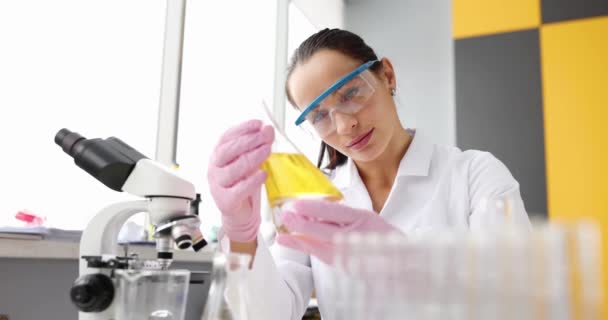 Une femme chimiste dans un laboratoire regarde un liquide jaune dans une fiole, au ralenti. Matériel de recherche médicale - Séquence, vidéo