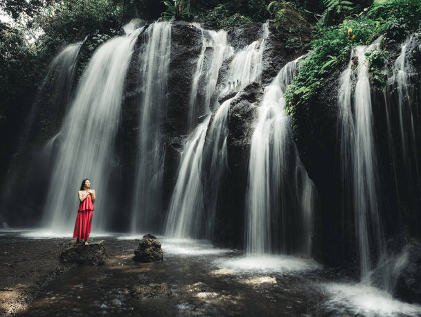 アジアの女性が滝の近くにいる. 自然と環境。 モーションウォーター。 トラベルライフスタイル。 長い赤いドレスを着ている若い女性. スリムボディ。 スペースをコピーする。 ウォーターフォールバリ。 モーション写真. - 写真・画像