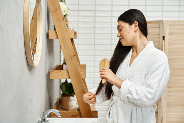 vue latérale de brunette et asiatique jeune femme en robe peigner ses cheveux bruns près miroir de salle de bain - Photo, image