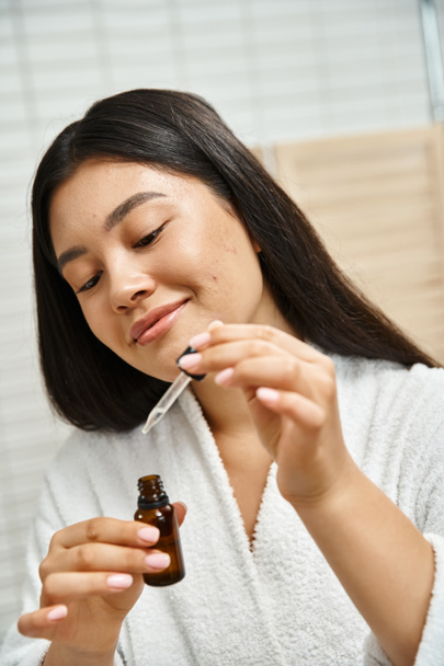 mulher alegre e asiática em roupão de banho segurando conta-gotas com soro facial para tratar acne no rosto - Foto, Imagem