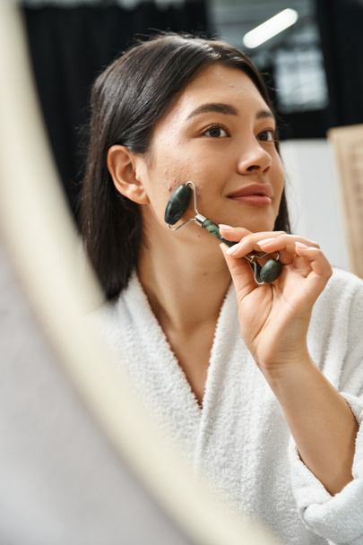 smiling asian woman with brunette hair using jade roller for a facial massage in bathroom, skin care - Photo, Image