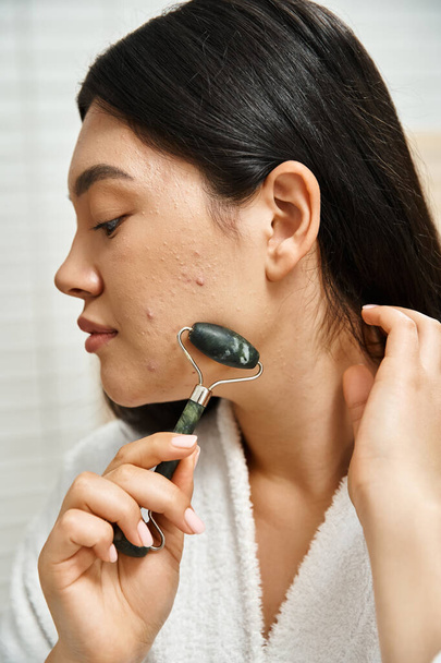 foto ravvicinata di giovane donna asiatica con acne che fa massaggio facciale con rullo di giada in bagno - Foto, immagini