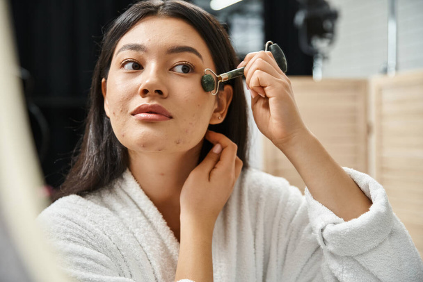 jonge aziatische vrouw met brunette haar met behulp van jade roller voor een gezichtsmassage in de badkamer, zelfverzorging - Foto, afbeelding