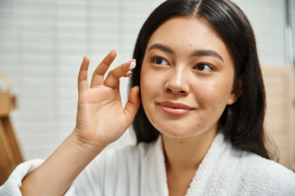 Nahaufnahme von jungen asiatischen Frau mit Akne Creme auf Gesicht und Blick auf Spiegel im Badezimmer - Foto, Bild