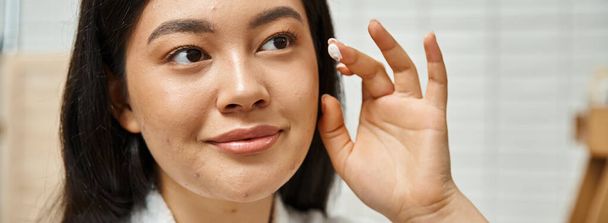 bannière de jeune femme asiatique avec de l'acné appliquer de la crème sur le visage et regarder miroir dans la salle de bain - Photo, image