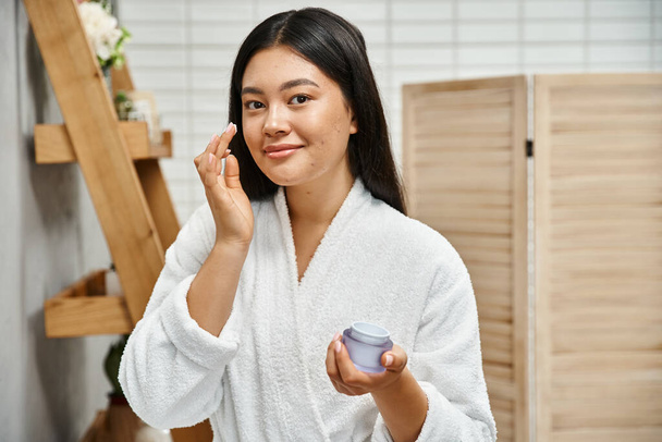 vrolijk aziatische vrouw met acne aanbrengen crème op gezicht en glimlachen terwijl kijken naar camera thuis - Foto, afbeelding