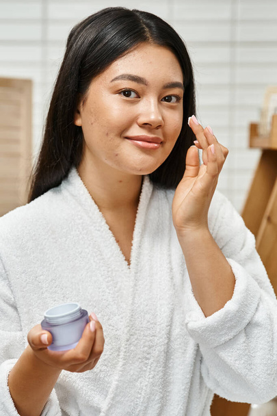 cheerful asian woman with acne applying cream on face and smiling while looking at camera, vertical - Photo, Image