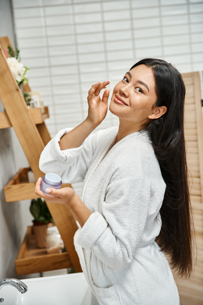 heureux et jolie asiatique femme avec acné application crème sur le visage et souriant tout en regardant caméra - Photo, image