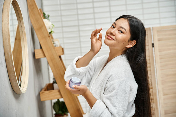 cheerful and pretty asian woman with acne applying cream on face and smiling while looking at camera - Photo, Image