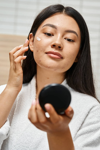 mancha tratamiento crema en la cara de la joven mujer asiática con acné mirando espejo compacto, vertical - Foto, imagen