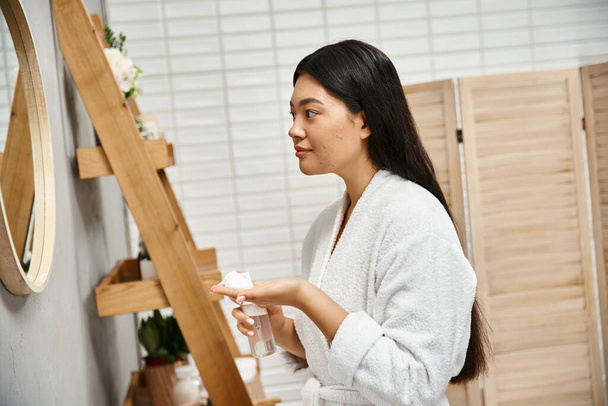 jeune femme asiatique avec des cheveux bruns se préparant à utiliser de la mousse nettoyante faciale dans la salle de bain moderne - Photo, image
