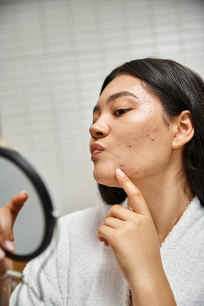 mladá asijská žena s brunetkou vlasy a pupínky zkoumání její tvář v zrcadle, kožní problémy - Fotografie, Obrázek