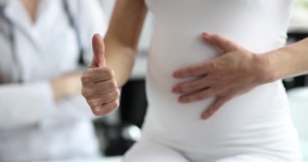 Een zwangere vrouw streelt haar buik en laat een duim zien, close-up. Vrouwenconsultatie, prenataal onderzoek - Video