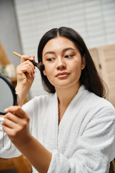 junge Asiatin mit brünetten Haaren schminkt sich mit Kosmetikpinsel über zu Akne neigende Haut - Foto, Bild