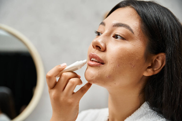 jovem e morena mulher asiática com acne aplicando bálsamo labial e olhando para o espelho no banheiro - Foto, Imagem