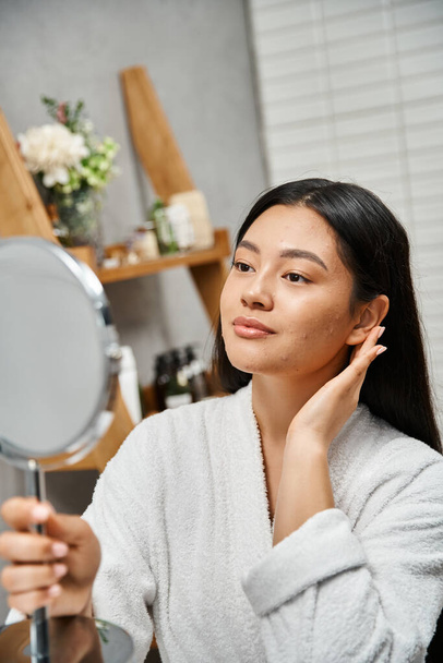 retrato de mujer joven asiática en bata tocando la piel propensa al acné y mirando el espejo en el baño - Foto, imagen