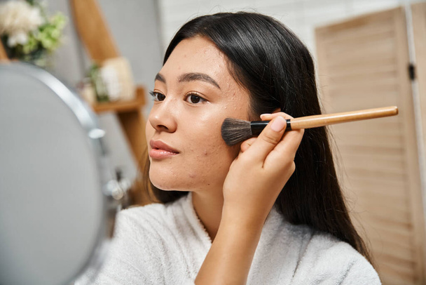 Esmer saçlı ve sivilceli genç Asyalı kadın yüz pudrası sürüyor ve aynaya bakıyor, deri sorunu var. - Fotoğraf, Görsel
